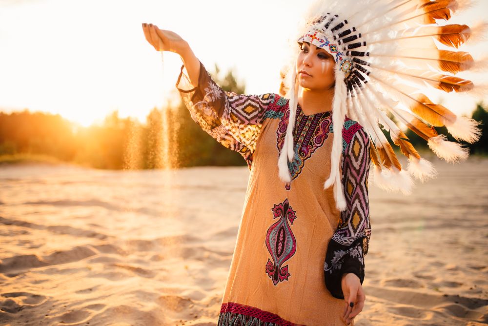 Beautiful woman in a suit of the American Indian play with sand on sunset. Young girl in headdress made of feathers of wild birds. Woman in suit of American Indian on sunset