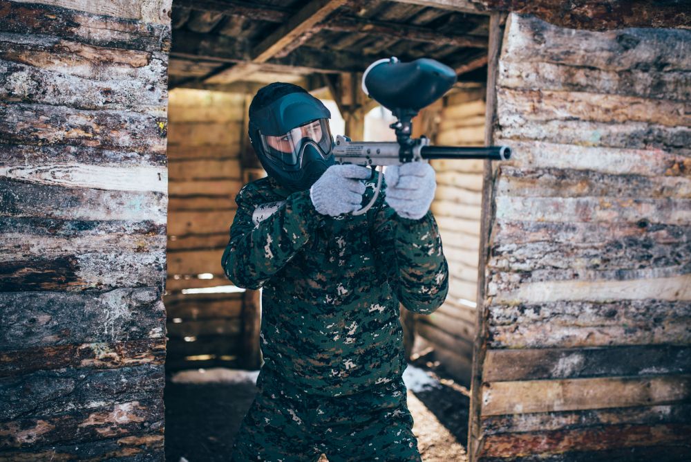 Male paintball player with marker gun in hands, front view, focus on weapon, winter battle. Extreme sport game, soldier fights in protection mask and uniform. Paintball player with marker gun, front view