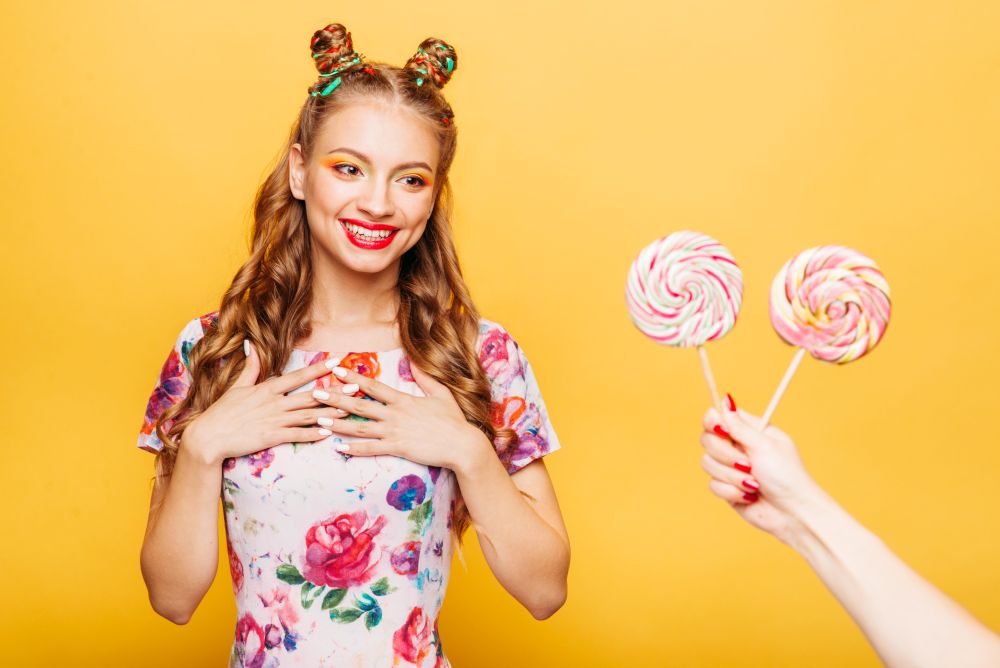 Beautiful young woman surprised somebody gives her lollypops. Bright girl with blonde curly hair. Stylish girl in summer colorful dress, yellow wall on background.. Woman surprised somebody gives her lollypops