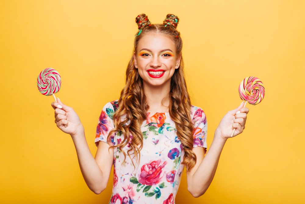 Young smiling lady holding two huge colorful lollypops. Amasing young woman fills happy and holding big candies in her hands. Stylish girl in summer dress, yellow wall on background.. Smiling lady holding two huge colorful lollypops