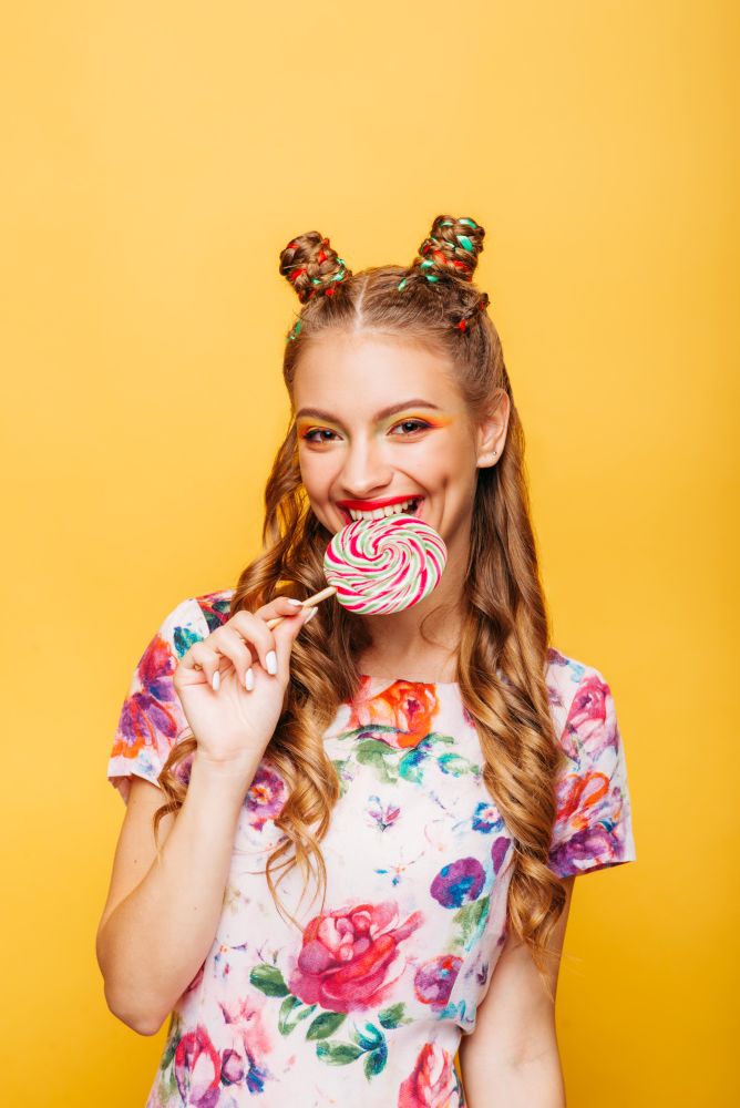 Beautiful young woman with playful look eating candy and smiling. Stylish girl with blonde curly hair. Portrait of attractive lady with big lollypop, yellow wall on background.. Woman with playful look eating candy