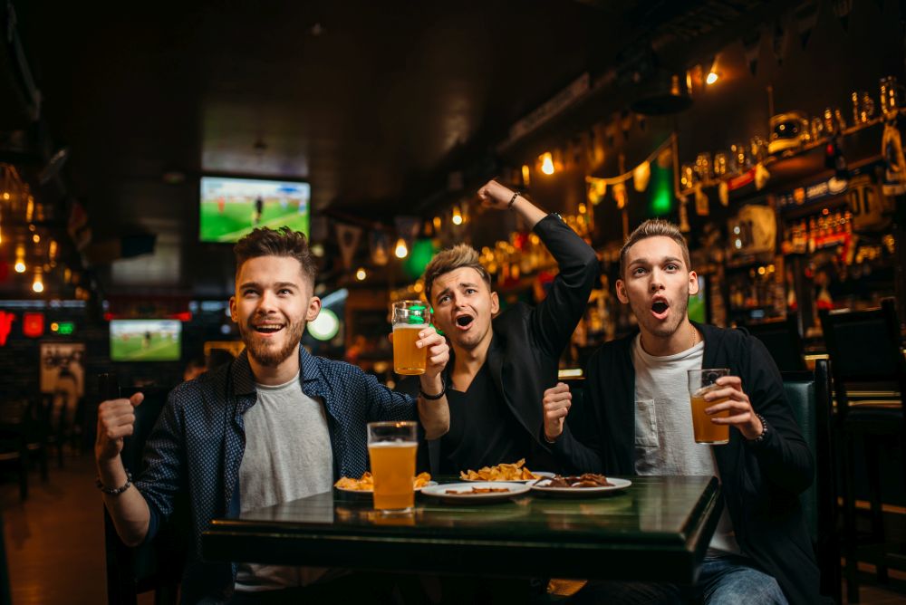 Three young men celebrates game victory in a sport bar, happy friendship of football fans. Men at the table with beer, crisps and crackers