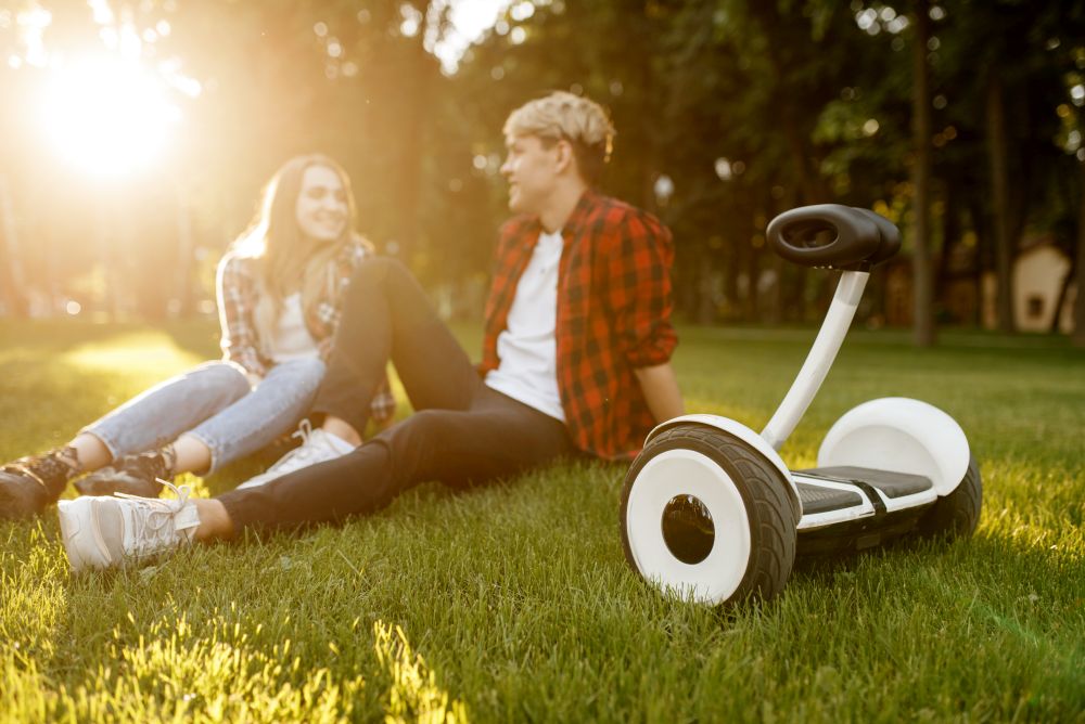Young couple sitting on the grass near gyro board in summer park. Outdoor recreation with electric gyroboard. Eco transport with balance technology. Young couple sitting on the grass near gyro board