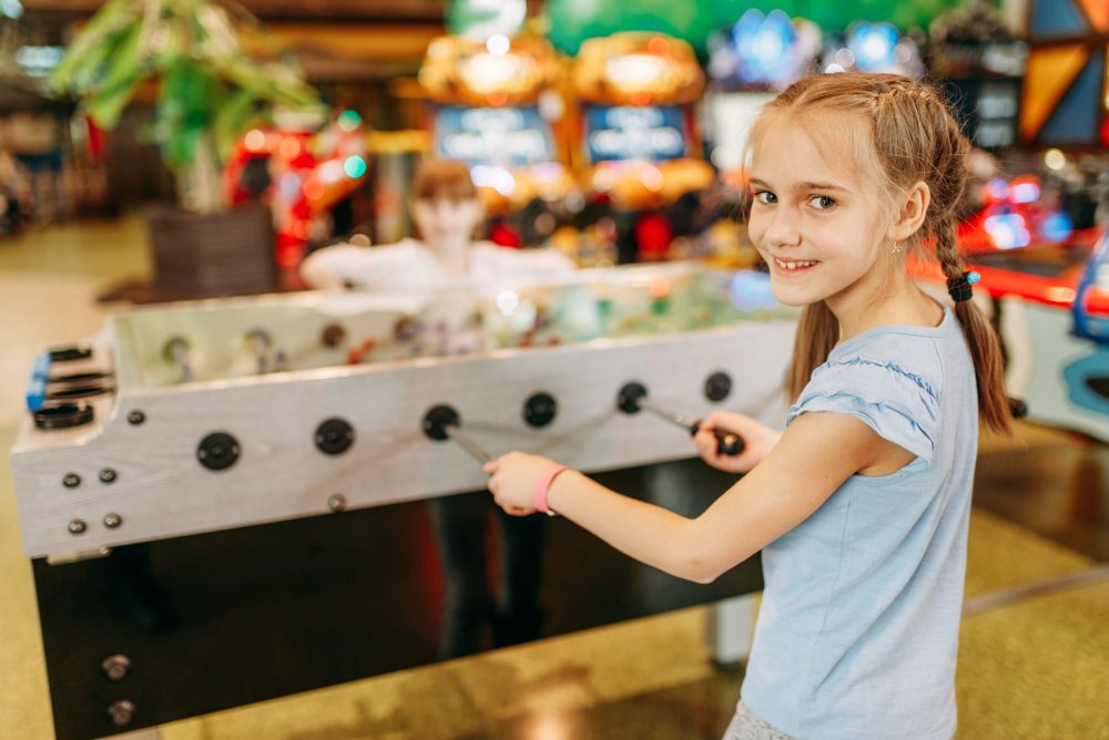 Happy girl at the table soccer in children game center. Smiling female child having fun on playground indoors. Kid plays on football machine in amusement centre. Happy girl at table soccer in children game center