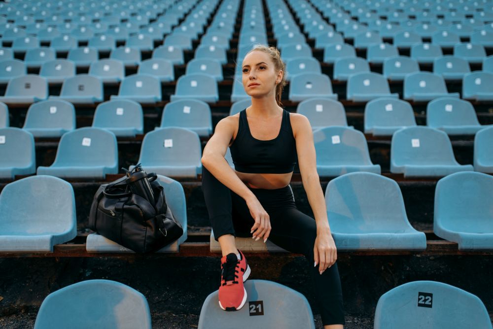 Female jogger in sportswear sitting on tribune, training on stadium. Woman doing stretching exercise before running on outdoor arena