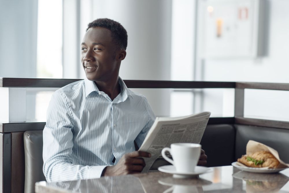 Young ebony businessman with newspaper having lunch in office cafe. Successful business person drinks coffee in food-court, black man in formal wear. Ebony businessman having lunch in office cafe