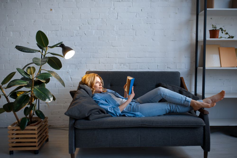 Young woman lying on cozy black couch and reading a book, living room in white tones on background. Attractive female person with magazine rest on sofa at home