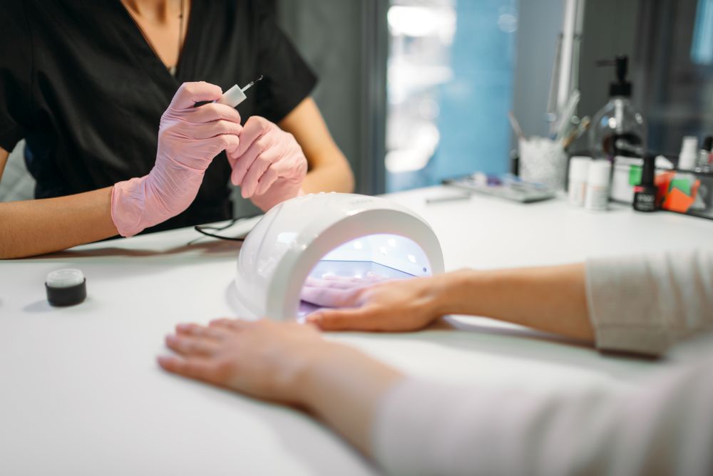 Female client dries gel varnish in the nail dryer, beautician in pink gloves on background, beauty salon. Manicurist doing hand care cosmetic procedure. Female client dries gel varnish in the nail dryer