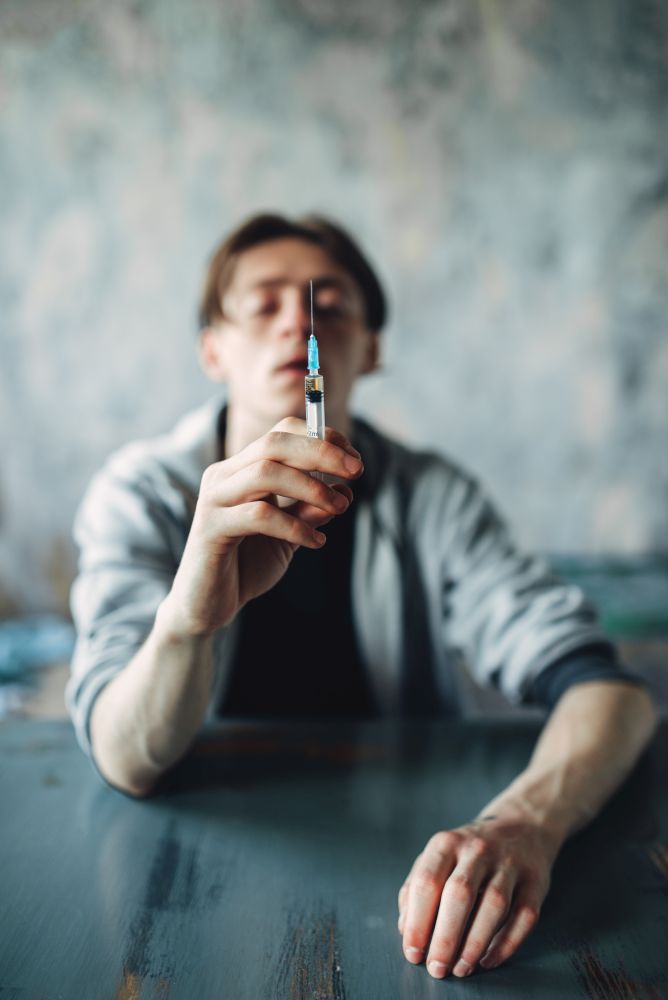 Male junkie sitting at the table with syringe in hand, grunge room interior on background. Drug addiction concept, addicted people. Junkie sitting at the table with syringe in hand