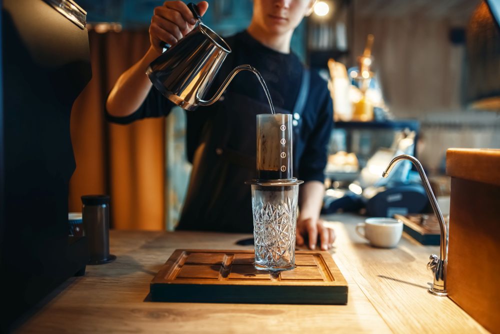 Male barista pours hot water from coffee pot into the glass, cafe counter and espresso machine on background. Barman works in cafeteria, bartender occupation. Barista pours water from coffee pot into the glass