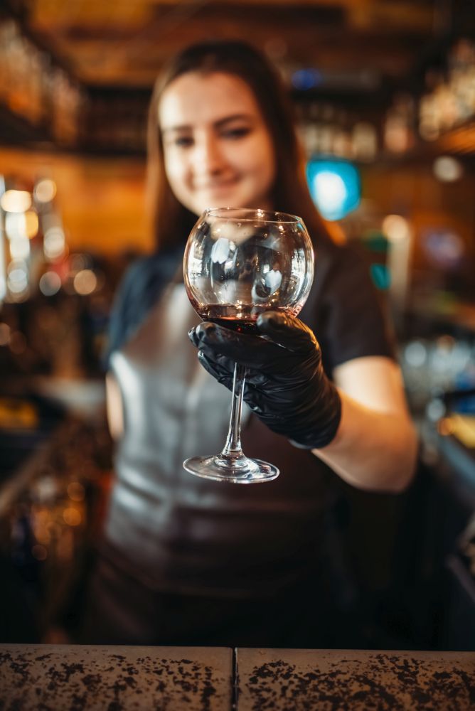 Female bartender in apron holds out a glass at the bar counter. Barman occupation, bartending. Female bartender in apron holds out a glass