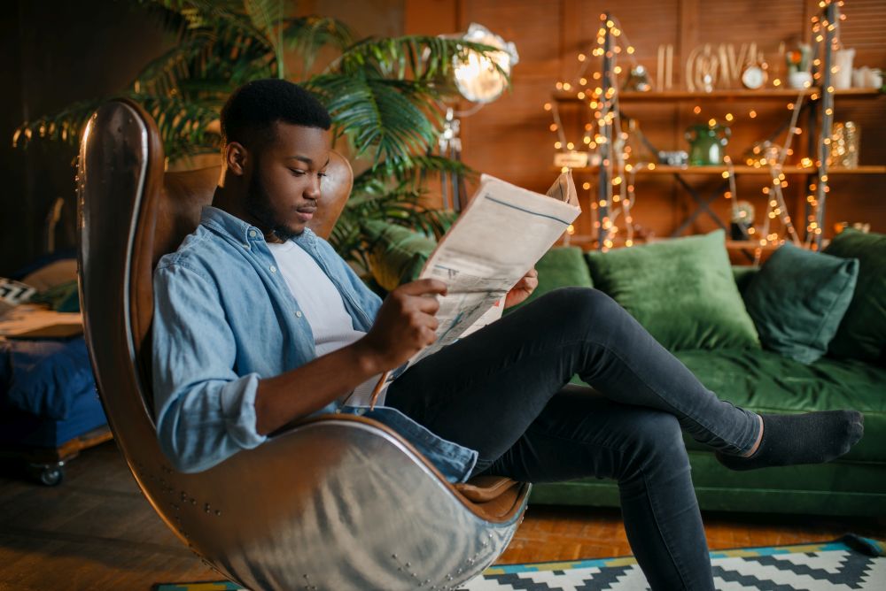 Black man reading newspaper in a comfortable leather chair in the living room, relaxation at home. Young african american male person in his house