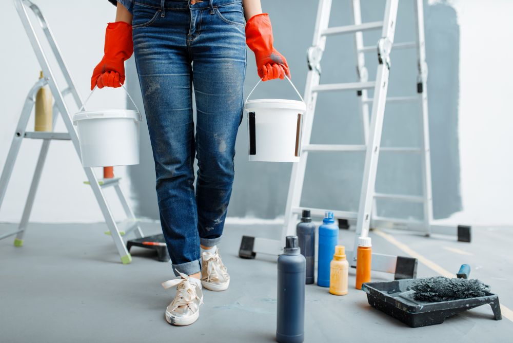 Female house painter holds pails with paints. Home repair, happy woman doing appartment renovation, room decoration renovating. Female house painter holds pails with paints