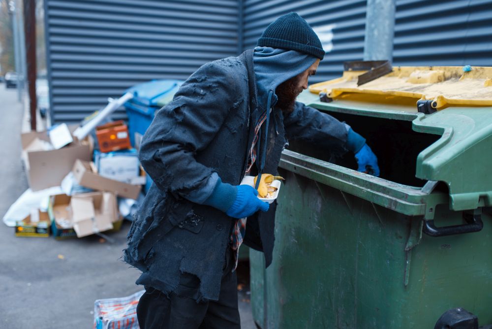 Male bearded beggar searching food in trashcan on city street. Poverty is a social problem, homelessness and loneliness, alcoholism and drunk addiction. Male bearded beggar searching food in trashcan