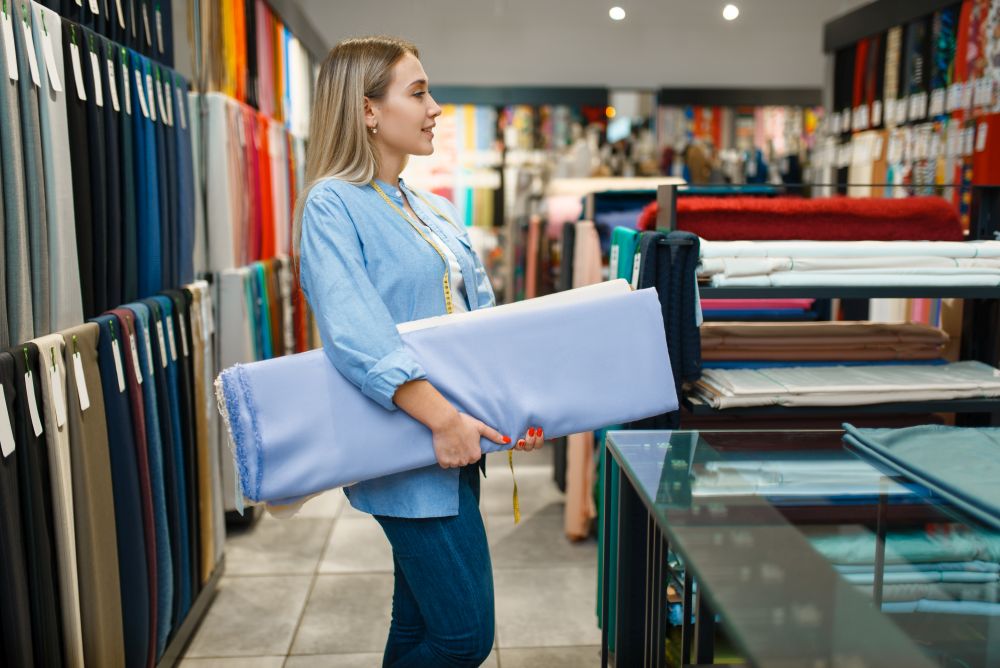 Seller holds stack of fabric in textile store. Shelf with cloth for sewing on background, clothing patterns choice in shop. Seller holds stack of fabric in textile store