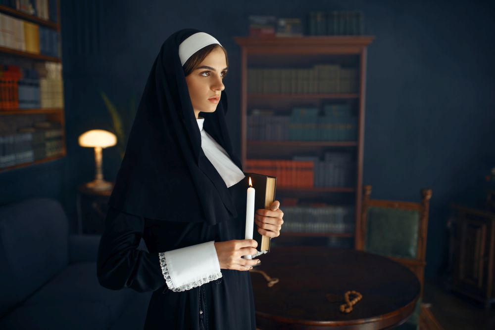 Young nun in a cassock holds a book and candle. The sister is preparing for prayer in the monastery, religion and faith, religious people. Young nun in a cassock holds a book and candle