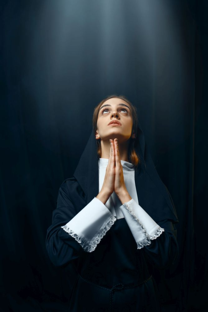 Young nun in a cassock prays on light over her head. The sister in the monastery, religion and faith, religious people. Nun in a cassock prays on light over her head