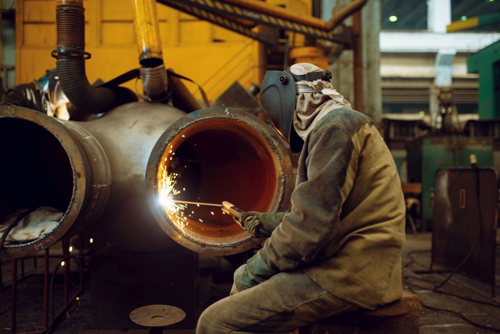 Welder in mask works with big metal pipe on factory, welding skill. Metalworking industry, industrial manufacturing of steel products. Welder works with metal on factory, welding skill