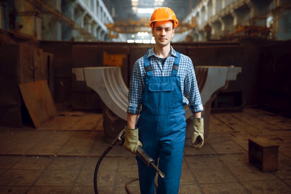 Male worker in uniform and helmet holds pneumatic jackhammer on factory. Metalworking industry, industrial manufacturing of steel products. Male worker holds pneumatic jackhammer on factory