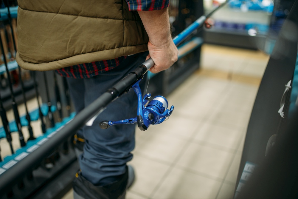 Male angler holds rod with reel in fishing shop. Equipment and tools for fish catching and hunting, accessory choice on showcase in store, spinnings and telescopes assortment. Male angler holds rod with reel in fishing shop