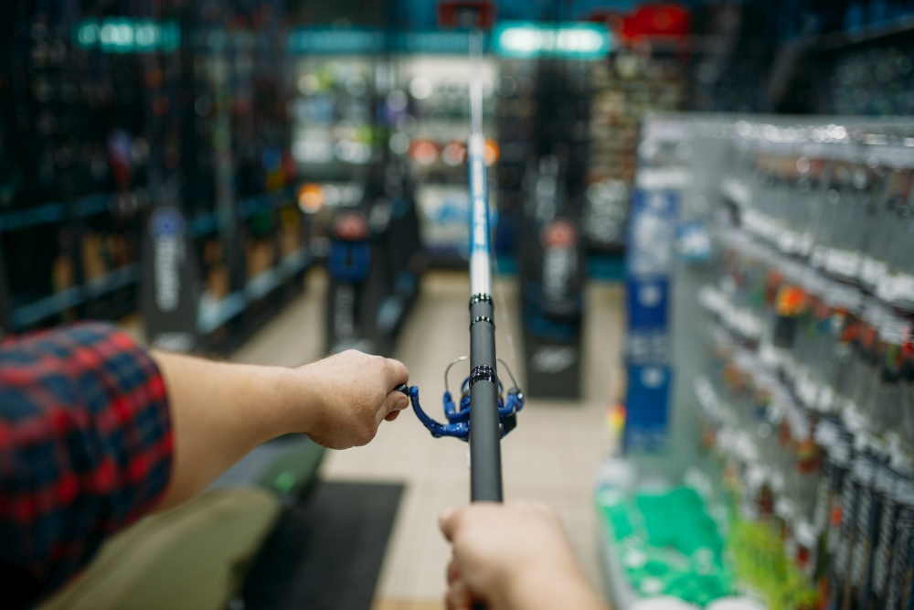 Male angler with rod in fishing shop, first-person view. Equipment and tools for fish catching and hunting, accessory choice on showcase in store, spinnings and telescopes assortment. Angler with rod in fishing shop, first-person view