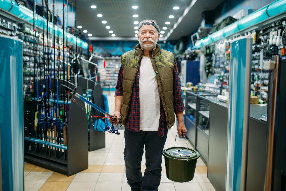 Fisherman with rod and plastic bucket in fishing shop. Male angler buying equipment and tools for fish catching and hunting, assortment on showcase in store. Fisherman with rod and bucket in fishing shop