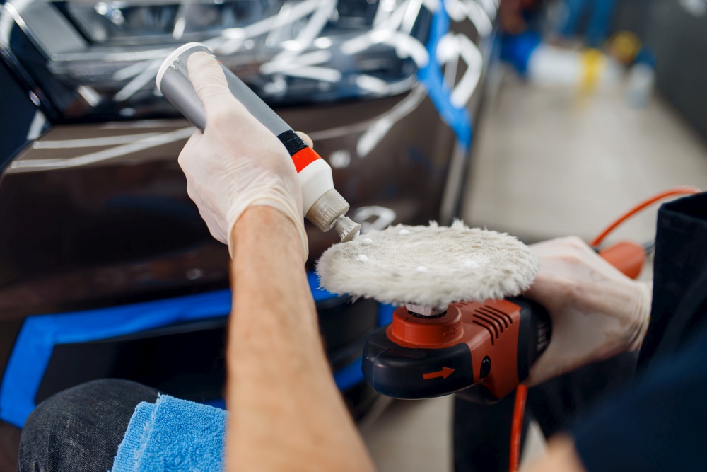 Male worker applies polish on polishing machine, car detailing. Preparation before installation of coating that protects the paint of automobile from scratches. Vehicle in garage, auto tuning. Male worker applies polish on polishing machine