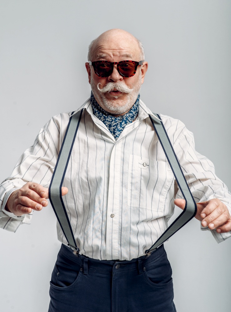 Fashionable elderly man in trousers with suspenders and sunglasses, grey background. Mature senior looking at camera in studio, dude. Fashionable elderly man, trousers with suspenders