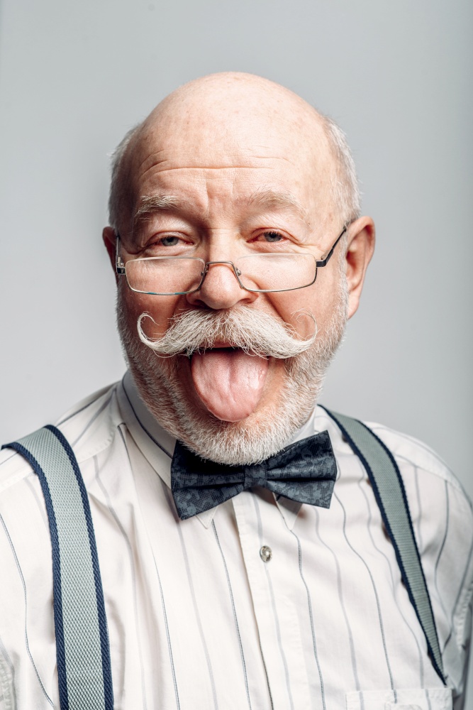 Portrait of elderly man in a bow tie and glasses showing his tongue, grey background. Mature senior looking at camera in studio. Portrait of elderly man showing his tongue