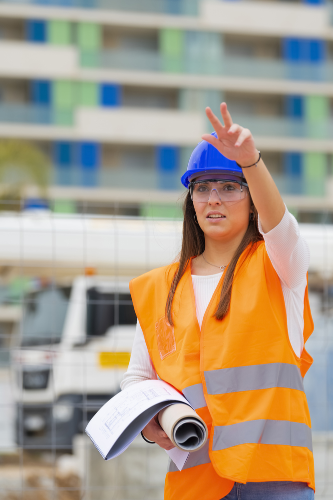 Closeup of a beautiful teenage architecture student holding blueprints and wearing safety helmet, glasses and vest raising her arm on an out of focus background. Work and apprenticeship concept.. Closeup of an architecture student with a raised arm