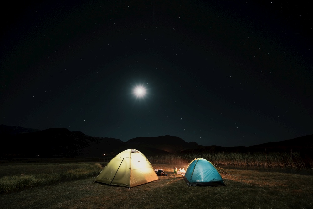 Camping in the mountains under the moon. A tent pitched up and glowing under the sky.. Tourist tents in camp among meadow in the night mountains