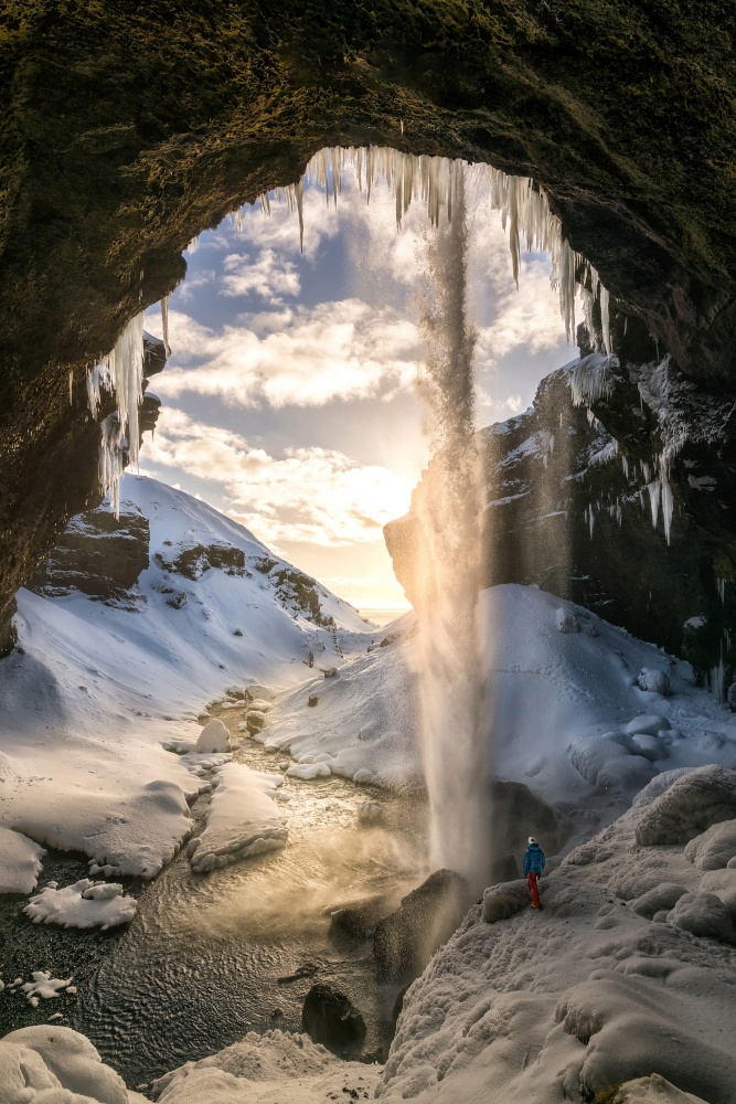 Spectacular waterfall in Iceland with guy looking at it at sunset time with water flowing through ceiling all snow covered