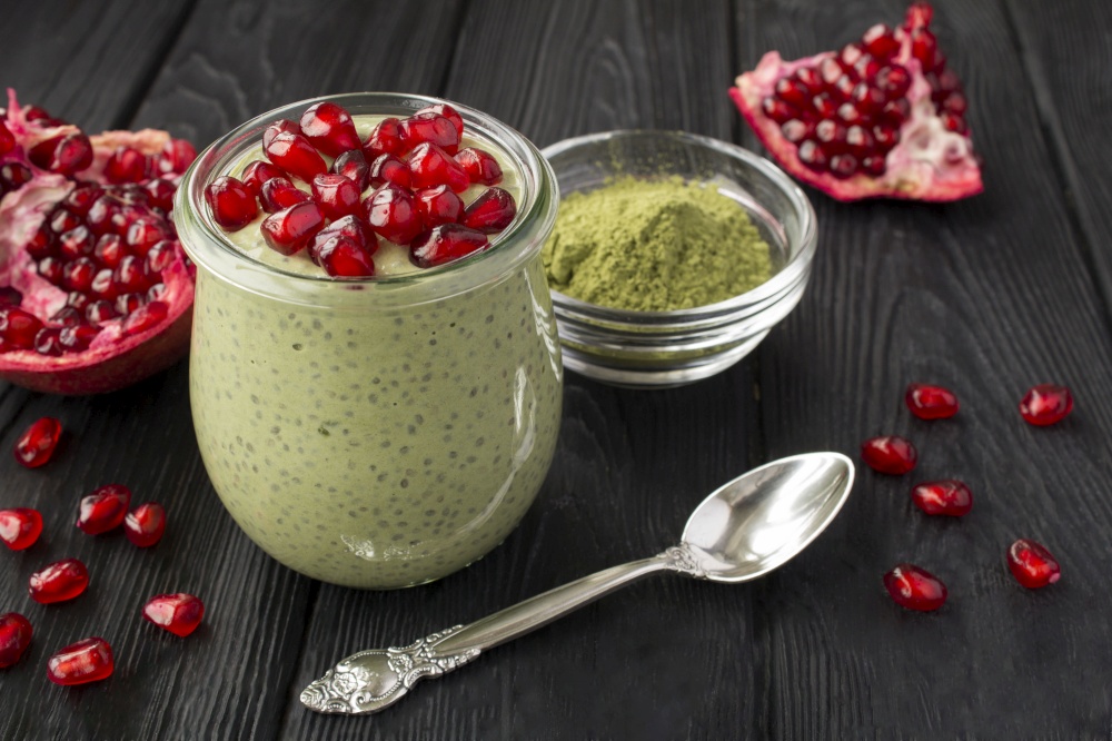Pudding with chia, matcha tea and pomegranate seeds in the glass jar on the black wooden background. Closeup.