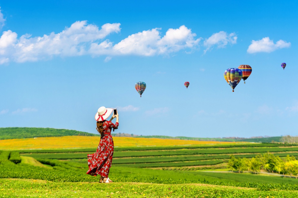 Woman standing on green grass and taking a photo to hot air balloons with smartphone.