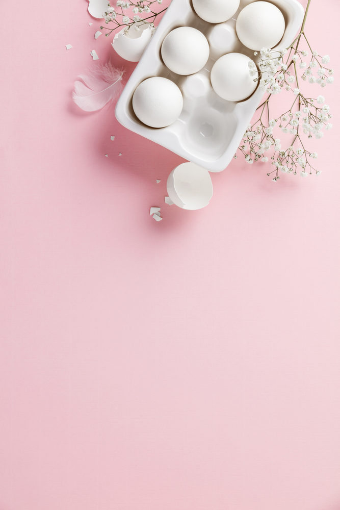 White eggs in white ceramic holder and flowers on pink background, flat lay, copy space
