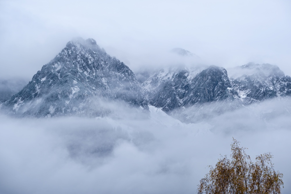 Overcast autumn alpine view with mountain silhoette fragments through fog and clouds. Climate, environment and weather concept sky background.
