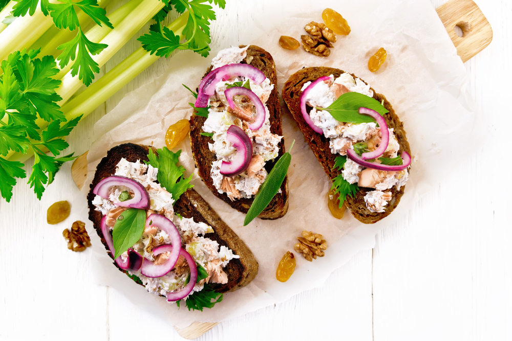 Salmon, petiole celery, raisins, walnuts, red onions and curd cheese salad on toasted bread with green lettuce on paper on a wooden board background from above