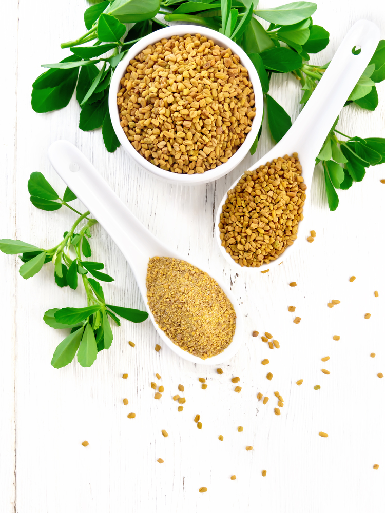 Ground fenugreek in a spoon, spice seeds in a bowl and spoon with green leaves on wooden board background from above
