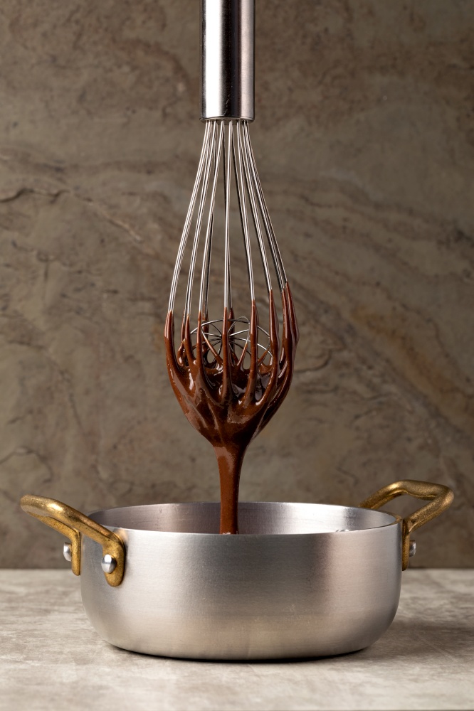 Liquid chocolate cream pouring from a whisk into a ladle. Dark background. . Liquid chocolate cream pouring from a whisk into a ladle.