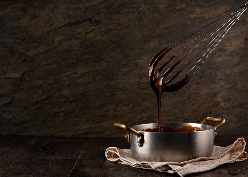 Liquid chocolate cream pouring from a whisk into a ladle. Dark background. . Liquid chocolate cream pouring from a whisk into a ladle.