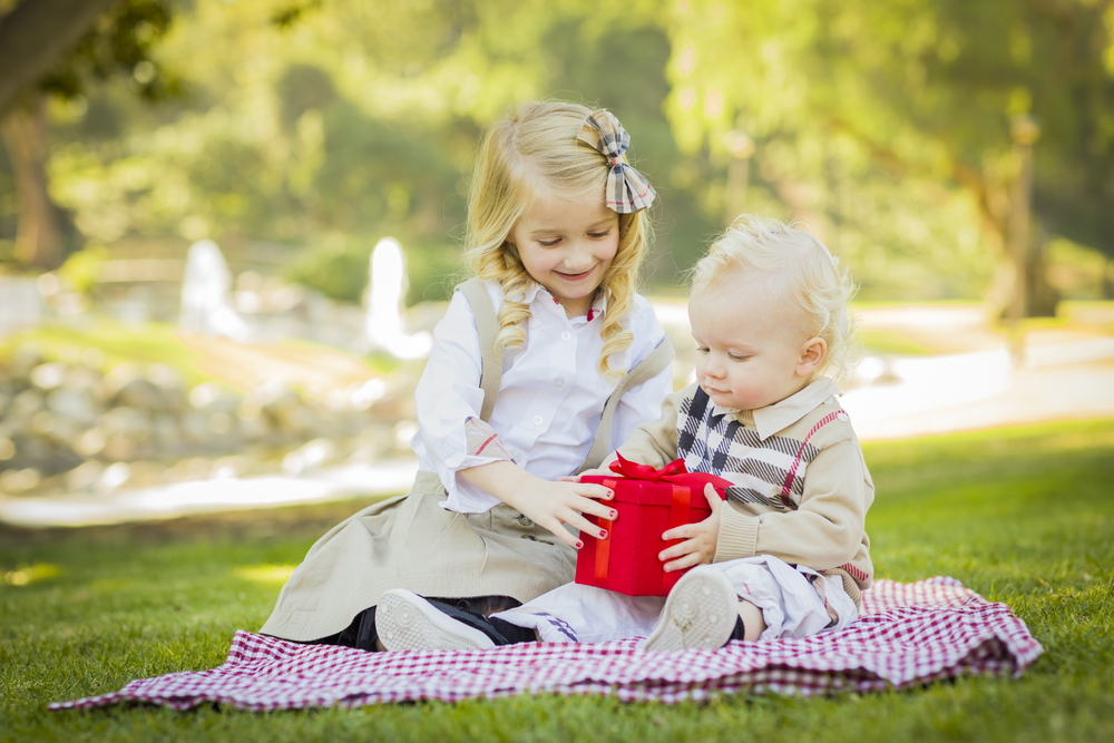 Sweet Little Girl Gives Her Baby Brother A Wrapped Gift on a Picnic Blanket Outdoors at the Park.